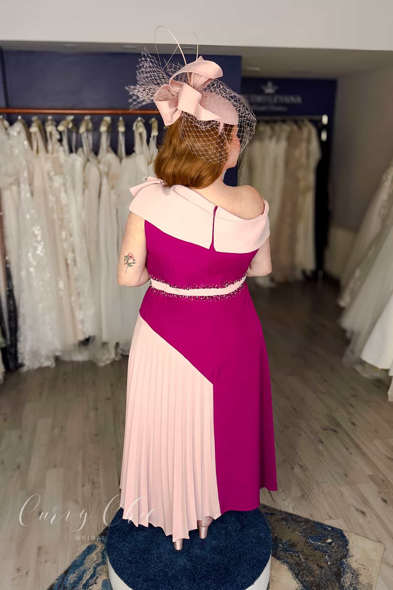 Plus size model wearing berry and blush asymmetric occasion dress with pleated skirt, back view, Curvy Chic Boutique Belfast