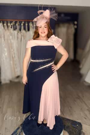 Plus size model wearing navy and blush off-the-shoulder occasion dress with rose detail and pleated panel, front view, Curvy Chic Boutique Belfast