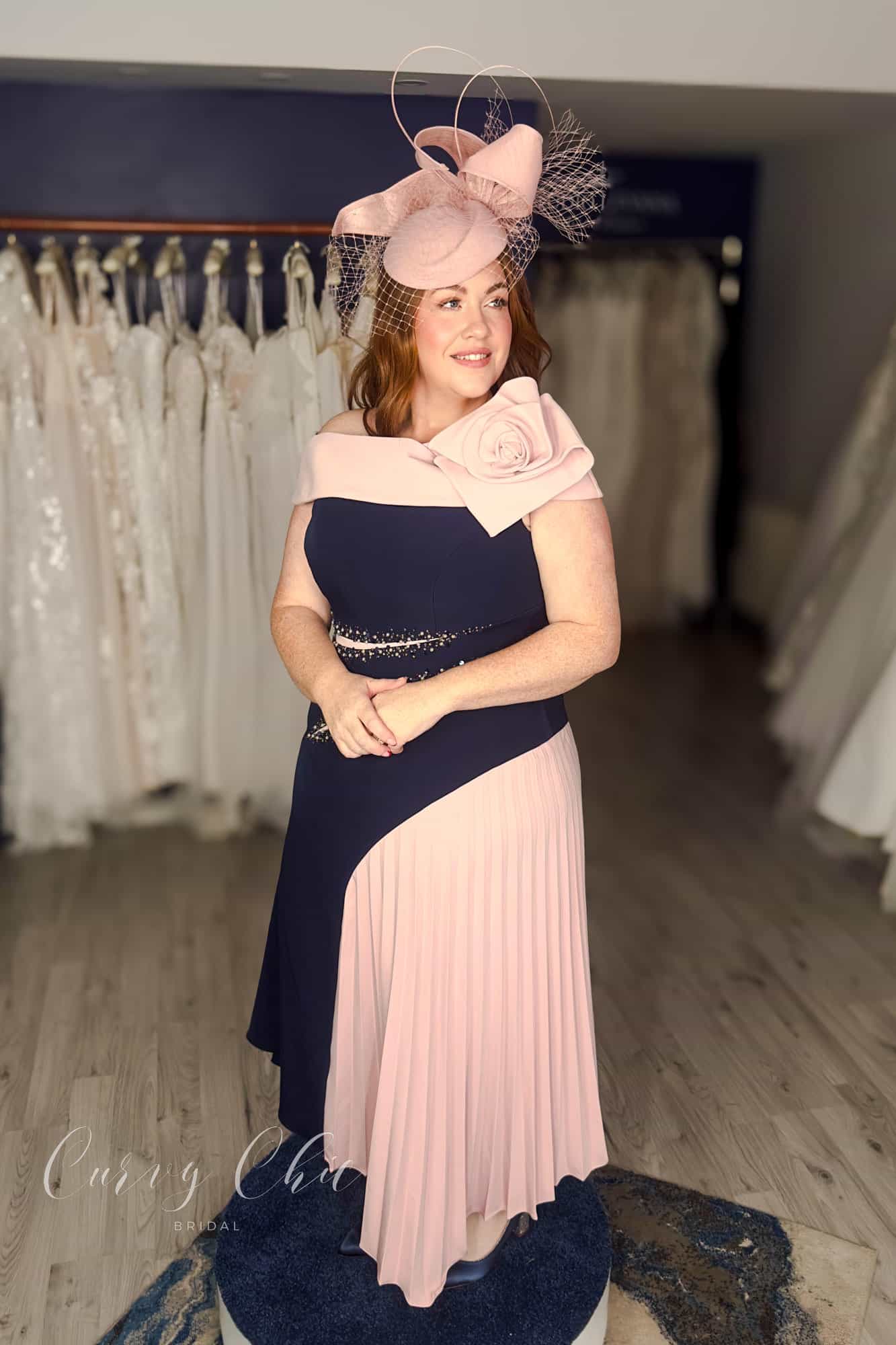 Side view of plus size model in navy and blush formal occasion dress with flowing pleated panel, Curvy Chic Boutique Belfast